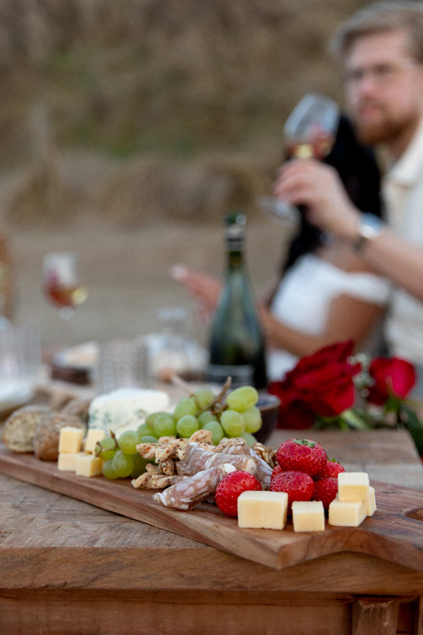 food board with cheese, strawberries, grapes, sausages and bread at picnic on the beach, with couple drinking wine in the background